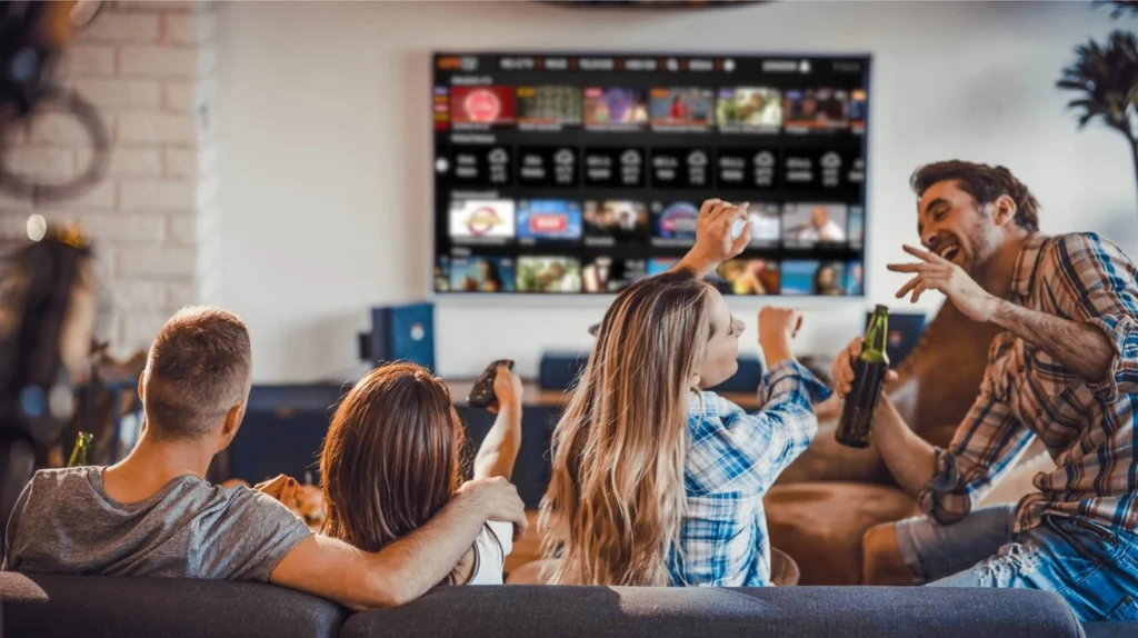 A group of friends sitting on a couch, enjoying entertainment on a large TV screen showing a streaming interface with multiple content thumbnails. The friends appear excited and engaged, with some holding beverages and raising their arms in celebration. The social scene depicts four people (three seated on the couch and one possibly sitting nearby) having an animated conversation or reaction to what's on the screen, illustrating how IPTV services can facilitate social entertainment experiences.