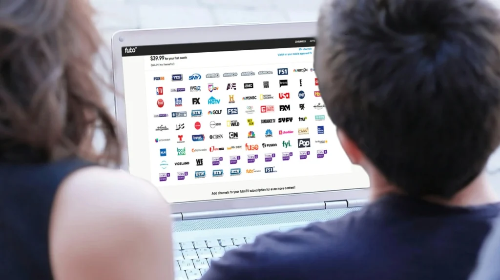 Two people looking at a laptop screen displaying the fuboTV streaming service interface. The screen shows "$39.99 for your first month" with a grid of numerous channel logos including FOX, YES, SNY, NBC, CBS, MSNBC, Fox News, FX, HGTV, and many other popular networks. The interface indicates there are over 275 channels available. This image demonstrates how IPTV/streaming services provide access to numerous traditional TV channels through a laptop, highlighting the portability and variety of content available through digital streaming platforms.