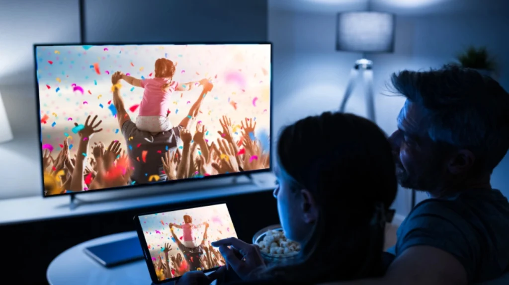 A couple watching content on both a TV and tablet simultaneously. The TV and tablet display the same vibrant scene showing a celebration with confetti and a child on someone's shoulders. They're enjoying what appears to be popcorn in a dimly lit room with blue ambient lighting. This image demonstrates the multi-device streaming capabilities of IPTV services, allowing viewers to enjoy synchronized content across different screens.