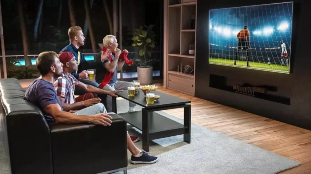 Four men watching an intense soccer/football match on a large wall-mounted TV in a modern living room during the evening. The TV shows a goalkeeper in an orange jersey ready to defend the goal. The men are visibly engaged and excited, with some standing and others sitting on a black leather couch. They have beers and snacks on the coffee table in front of them. This image captures the social aspect of watching live sports through IPTV services, bringing friends together for immersive viewing experiences.