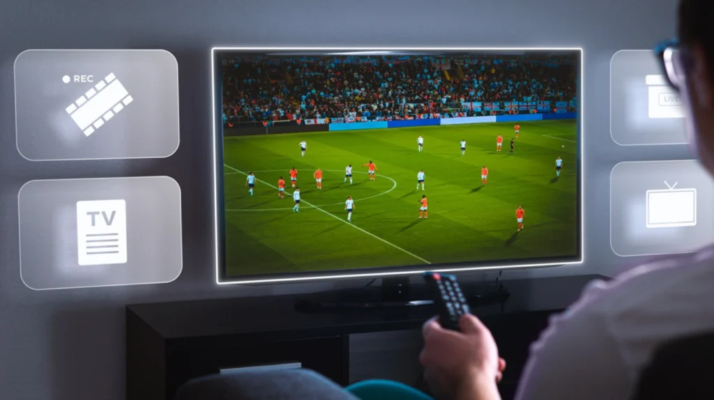 A TV displaying a live soccer/football match with players in white and orange/red jerseys on a green field with a crowded stadium in the background. Around the TV screen are interface icons including "REC" (recording), "TV" (guide), and "LIVE" icons, highlighting the DVR and live sports streaming capabilities of IPTV services. A person's hand is visible holding a remote control in the foreground, demonstrating the interactive nature of watching live sports through IPTV platforms